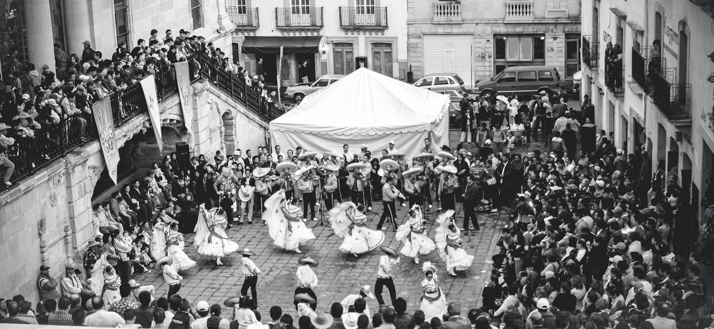 Un grupo de bailarines folclóricos y una banda de mariachis animan a una multitud en una plaza de la ciudad.